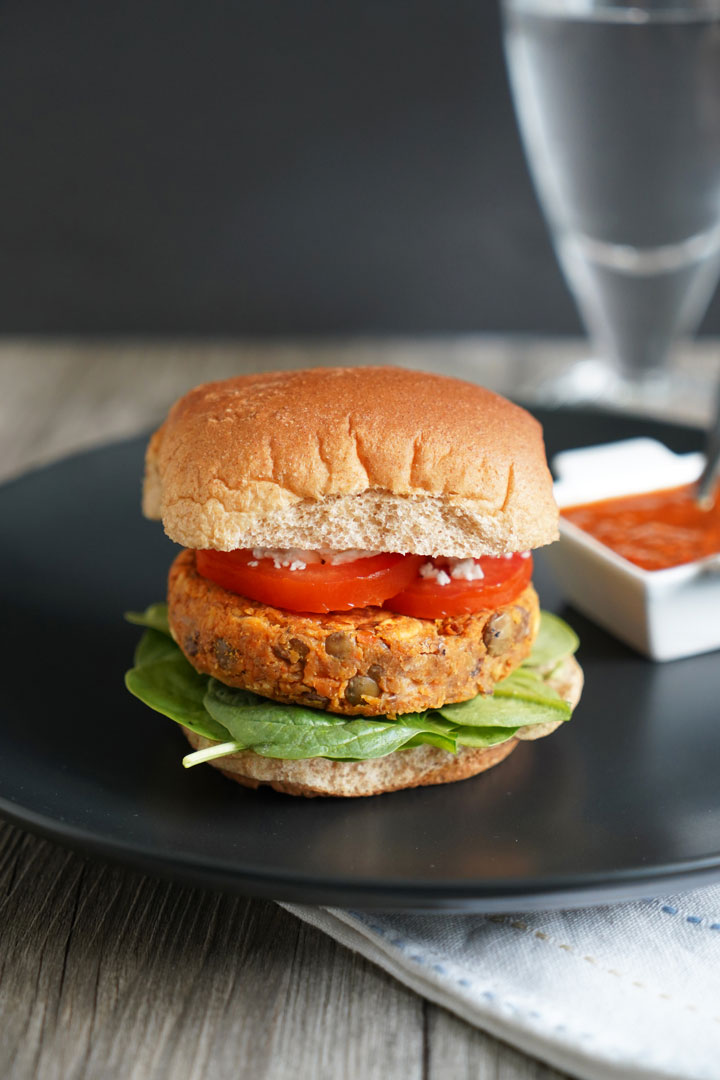 Lentil burger in whole wheat patty with tomato, lettuce on a black plate with harissa dressing on the side.