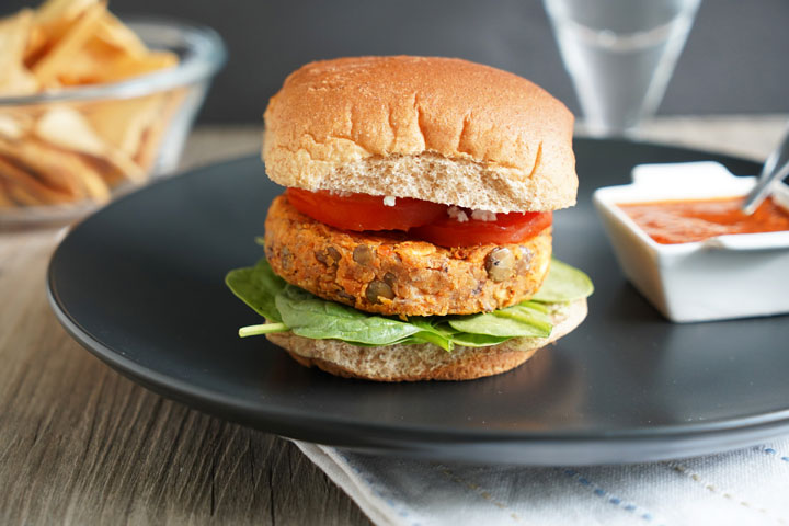 Lentil burger in whole wheat patty with tomato, lettuce on a black plate with harissa dressing on the side.