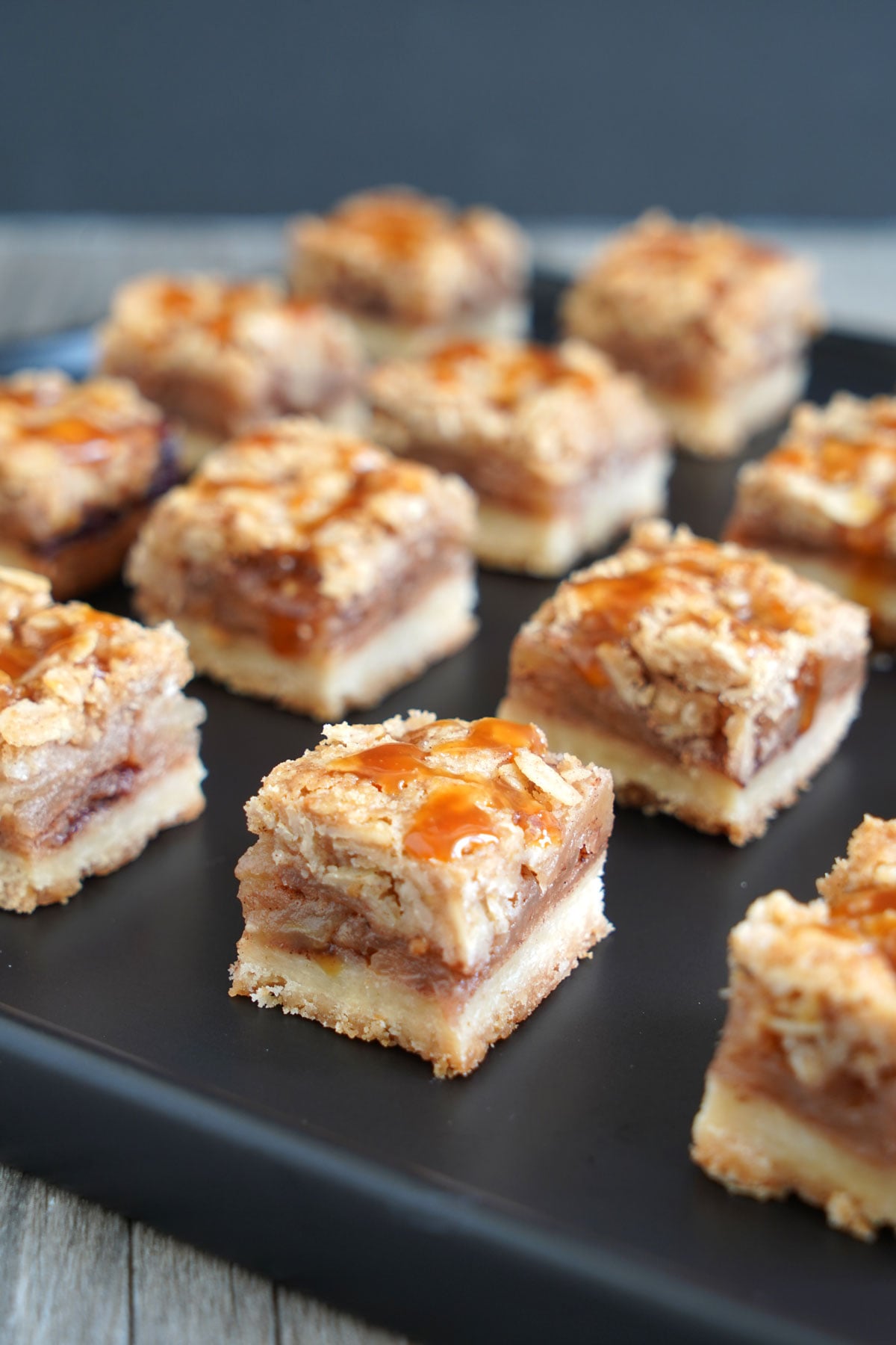 Bite-size salted caramel apple pie bars arranged in neat rows on a black serving tray.