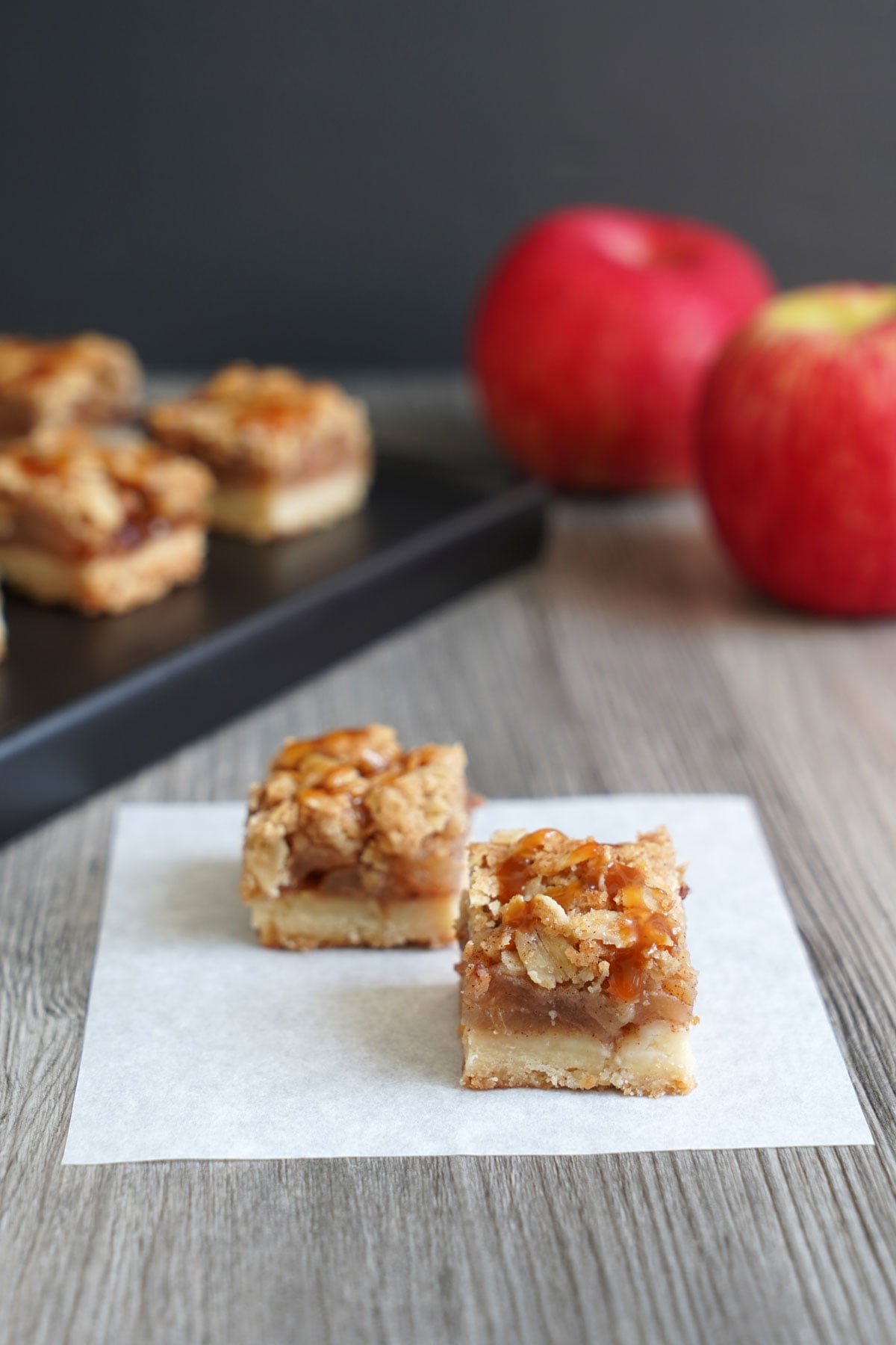Two apple pie bar squares on parchment paper with more bars and fresh apples in the background.