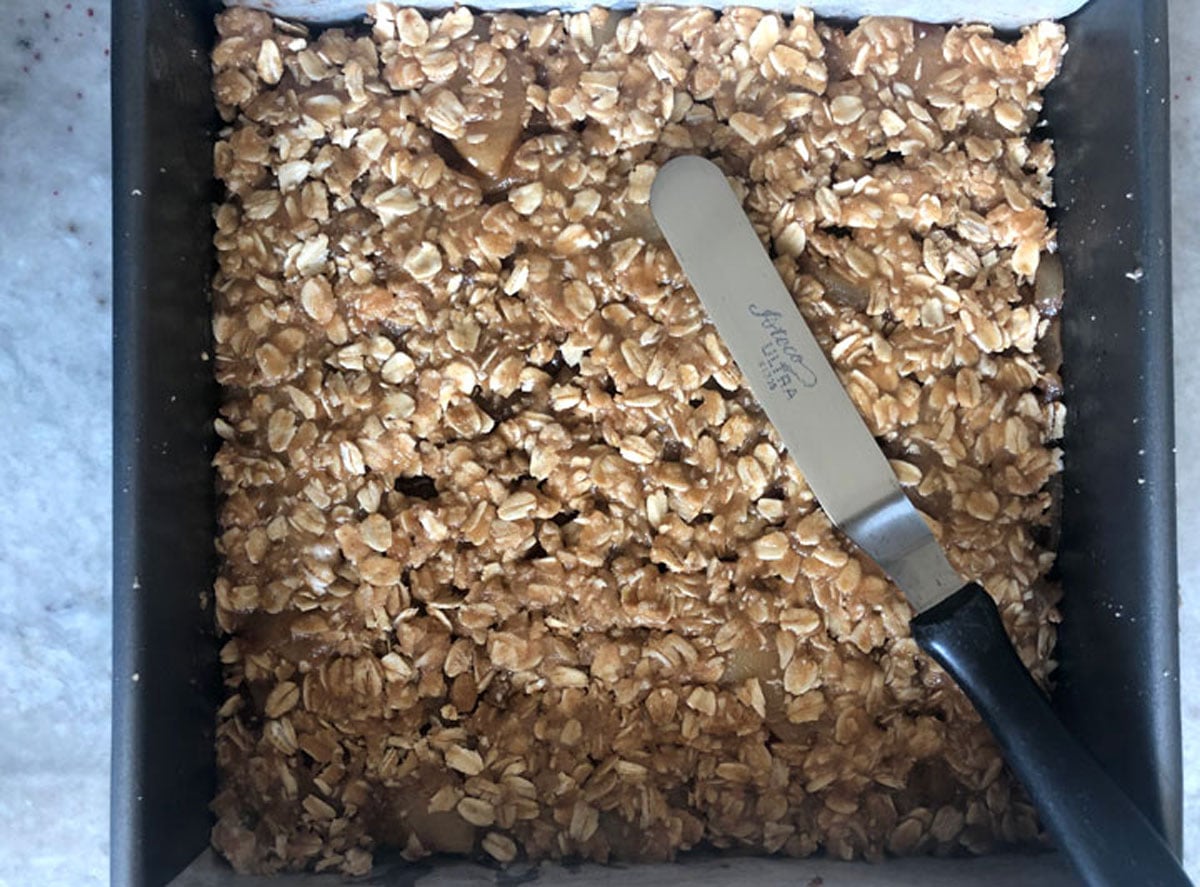 Streusel-topped apple pie bars smoothed flat in a parchment-lined pan with an offset spatula before baking.