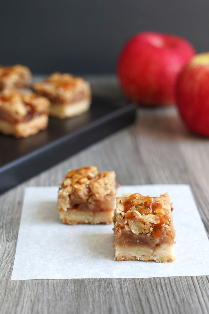 Two bite sized apple pie bars on a piece of parchment with more bars and apples in the background.