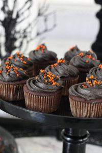 Chocolate cupcakes frosted with black frosting and orange and black sprinkles with halloween themed background.