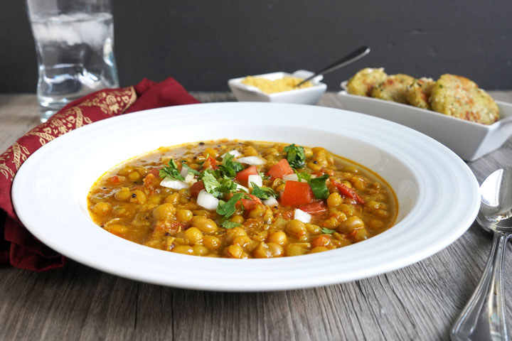 Ragda garnished with tomatoes, cilantro, and onion in a white plate. Patties and sev in background.