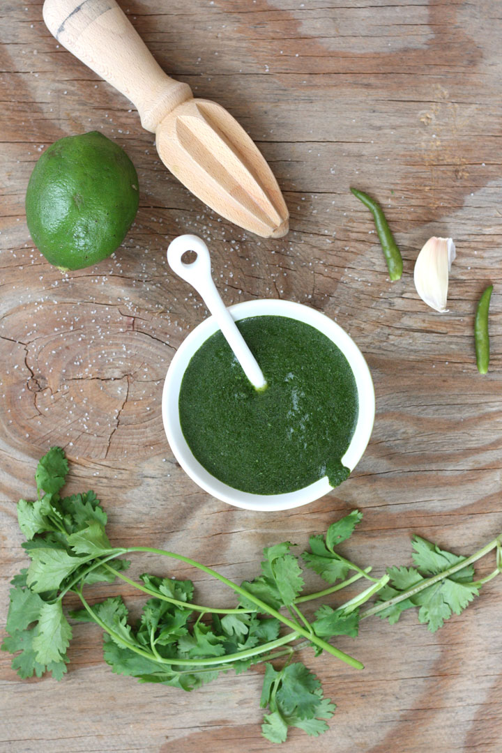 Cilantro chutney shown in small white bowl with limes, chilis and fresh garlic and cilantro around it. 