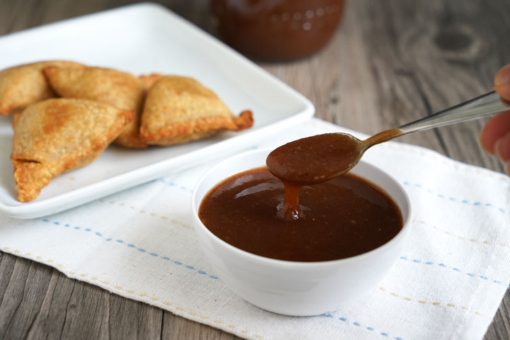 Tamarind date chutney being spooned from a white bowl with plate of samosas in background.