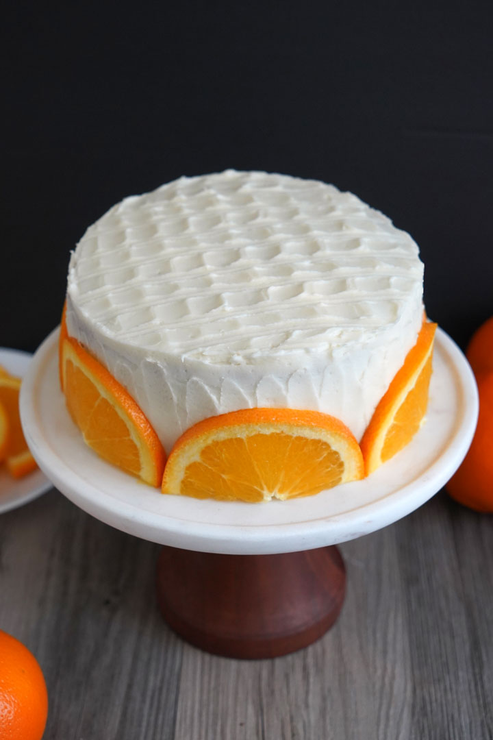 Orange cake with cream cheese frosting and fresh orange slices shown on a wood pedestal. 