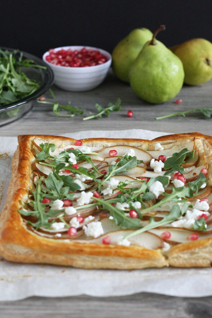 Pears baked on puff pastry with arugula, pomegranate, and goat cheese sprinkled on top. Pears in background.