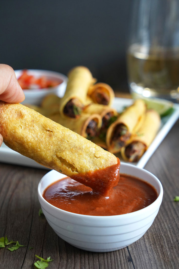 Taquito being dipped in enchilada sauce.