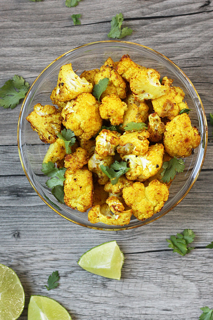 Roasted Cauliflower with turmeric in a clear glass bowl with cilantro and lime on the sides.