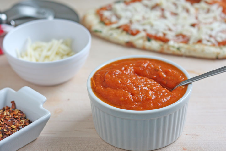 Pizza sauce in a bowl with pizza and cheese in background.