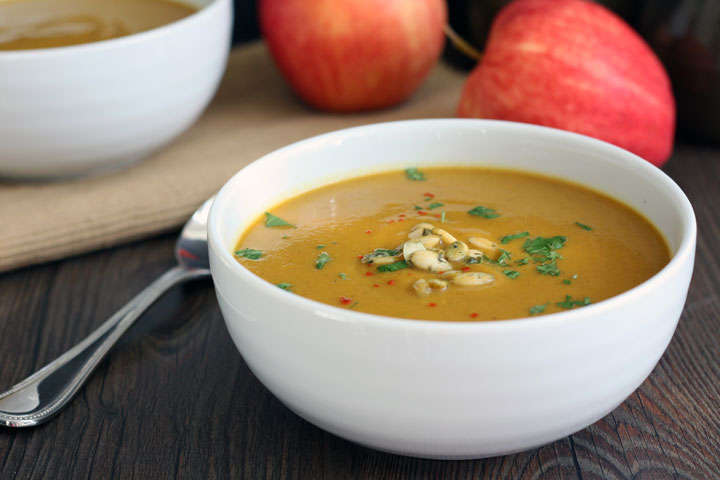 Soup in white bowl with spoon next it and apples in background.