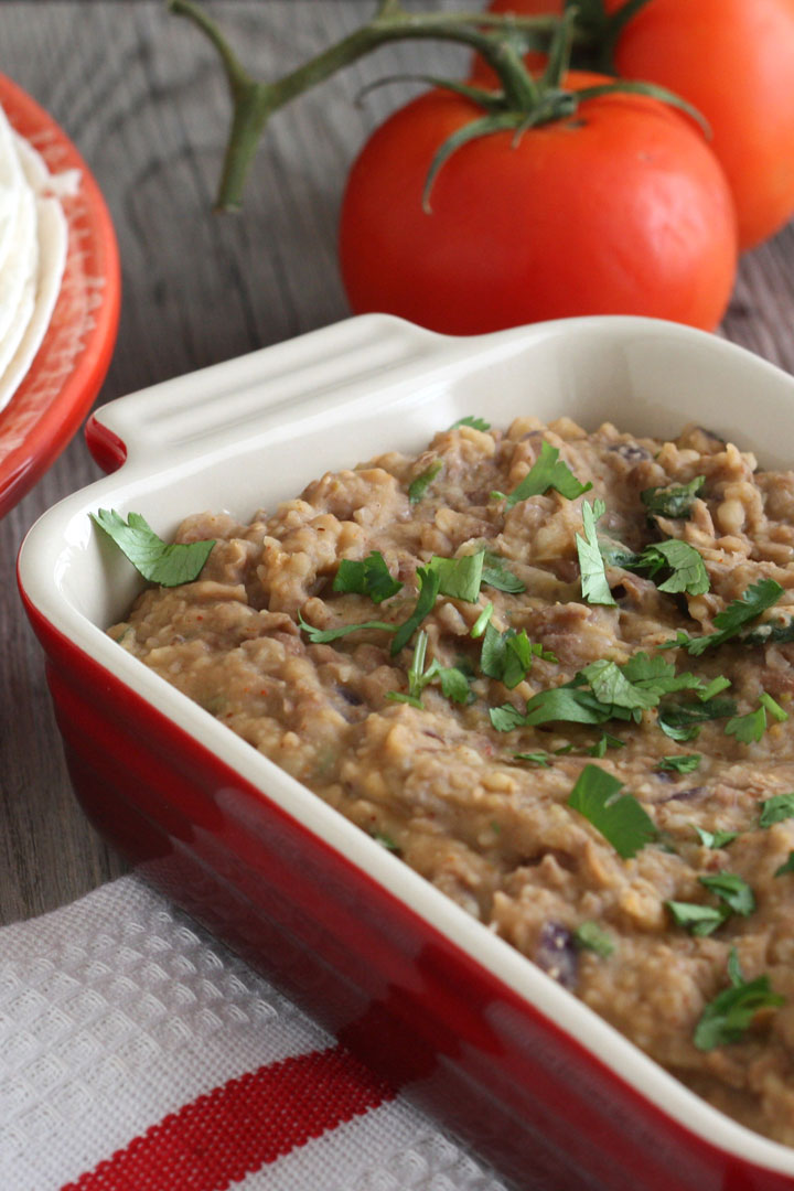 Close up view of refried beans in red serving dish with tomatoes in background.