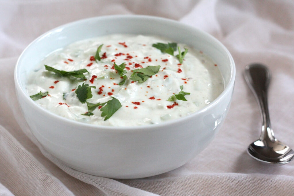 Cucumber raita in white bowl with spoon on the side.