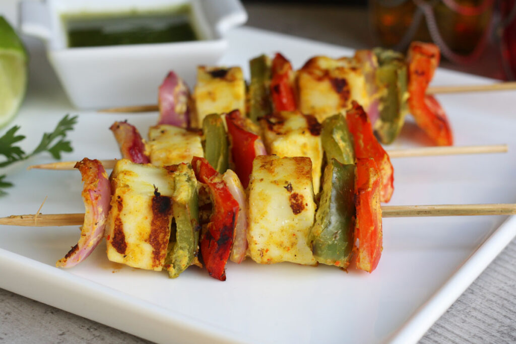 Finished paneer tikka on skewers in white plate with side of chutney.