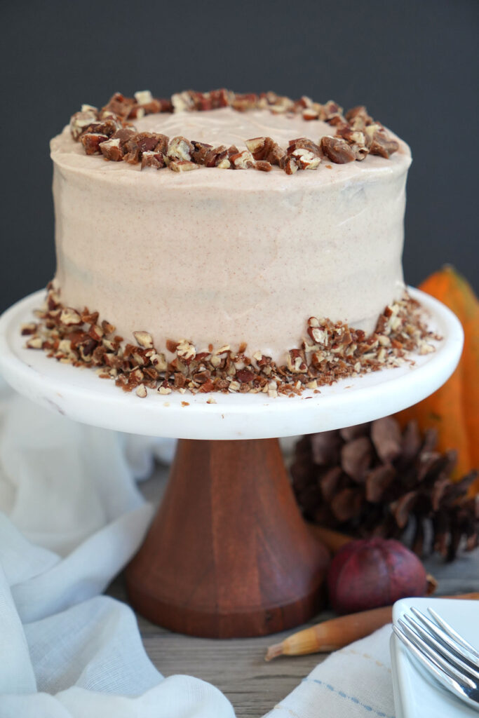 Sweet potato cake decorated with candied pecans on wooden and marble cake stand with acorns and squash in background.