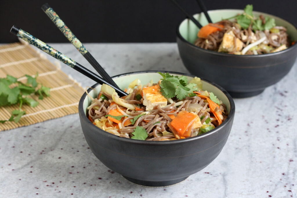 Sesame noodles in two bowls with chopsticks. Cilantro garnish on the side.