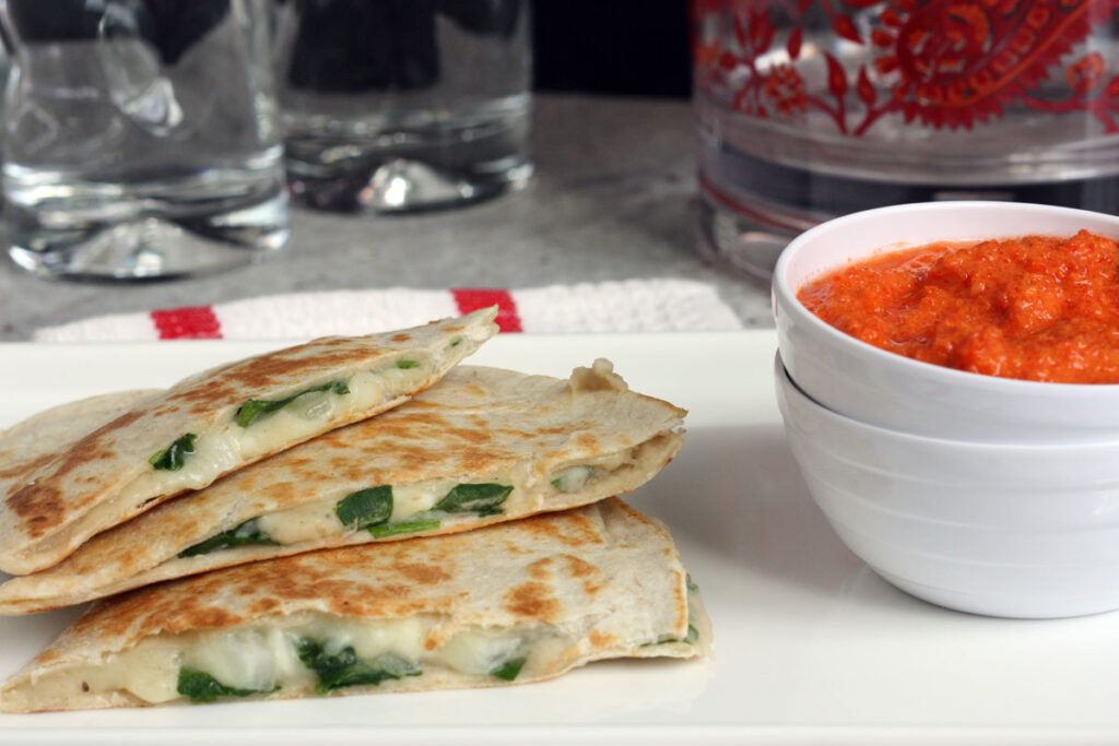 Three quesadillas in a plate next to a bowl of roasted red pepper dip. 