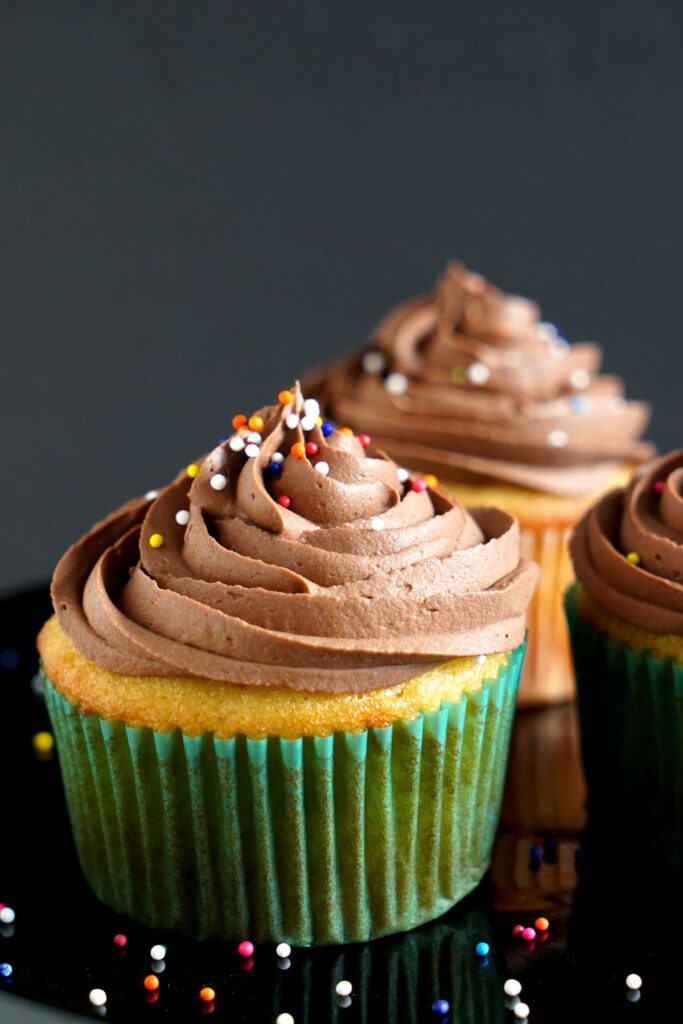 Close up of a vanilla cupcake in green wrapper frosted with fudge buttercream and topped with colorful round sprinkles. 