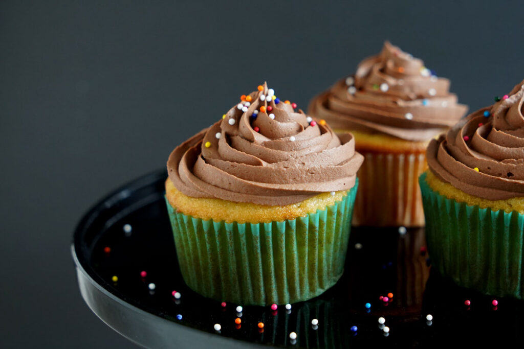 Chocolate frosted vanilla cupcakes on a black platter with sprinkles everywhere.