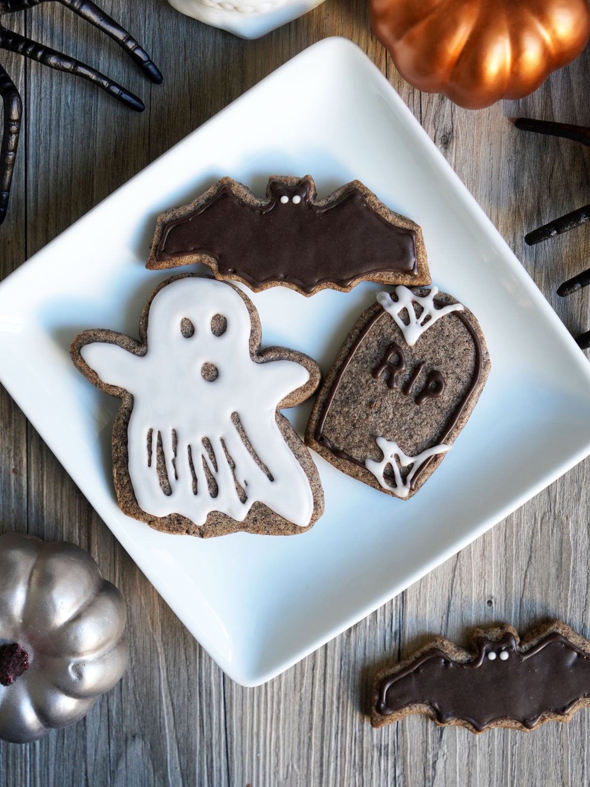 Halloween sugar cookies shaped like a bat, ghost, and tombstone on a white plate shown from above.