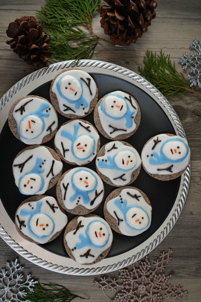 Melted snowman cookies in round black platter surrounded by snowflakes and pine cones.