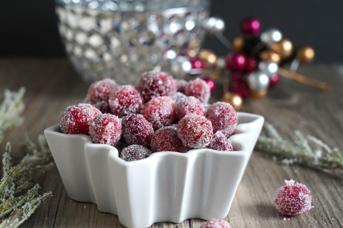 Sugared cranberries in white bowl with sugared rosemary on the side and festive decor in background.