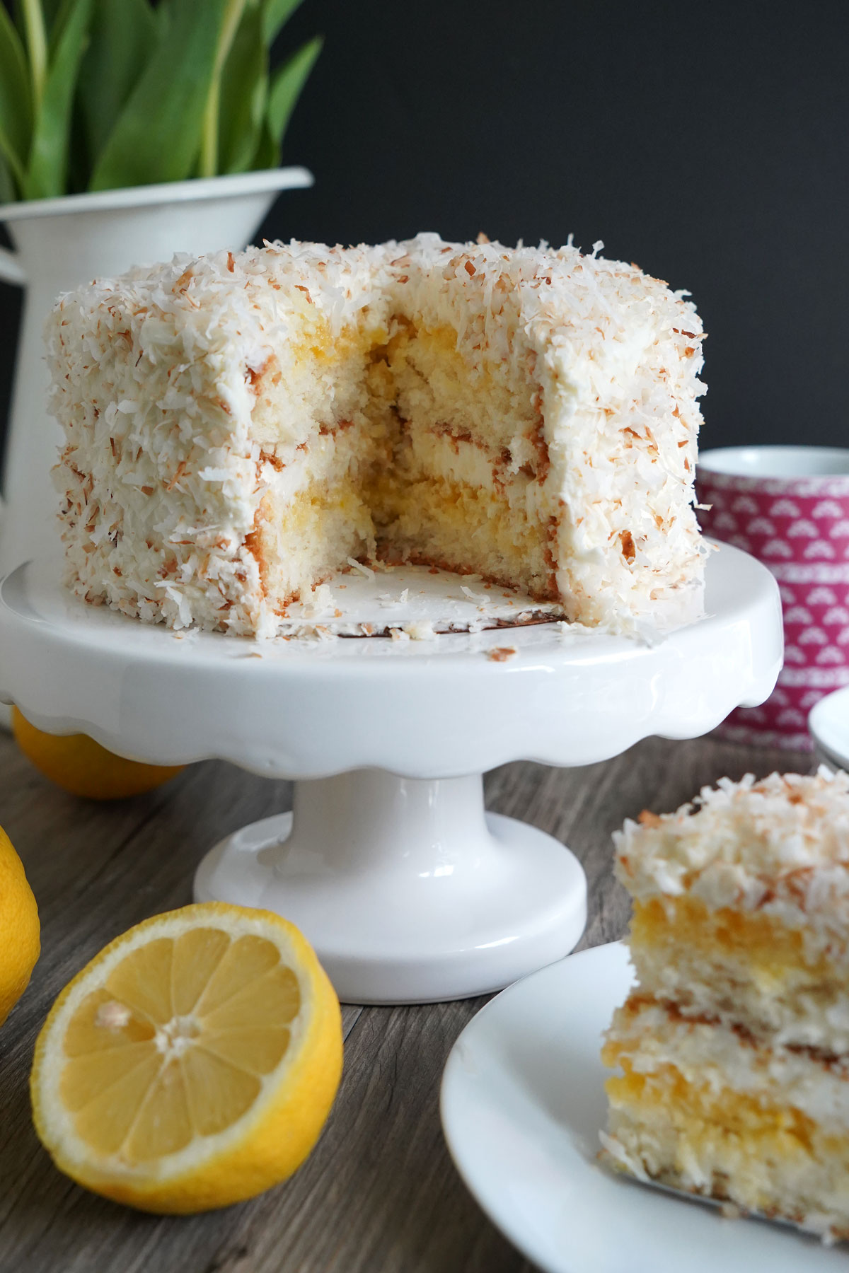 Lemon coconut cake cut open on a white pedestal. Cake slice and lemons in foreground.