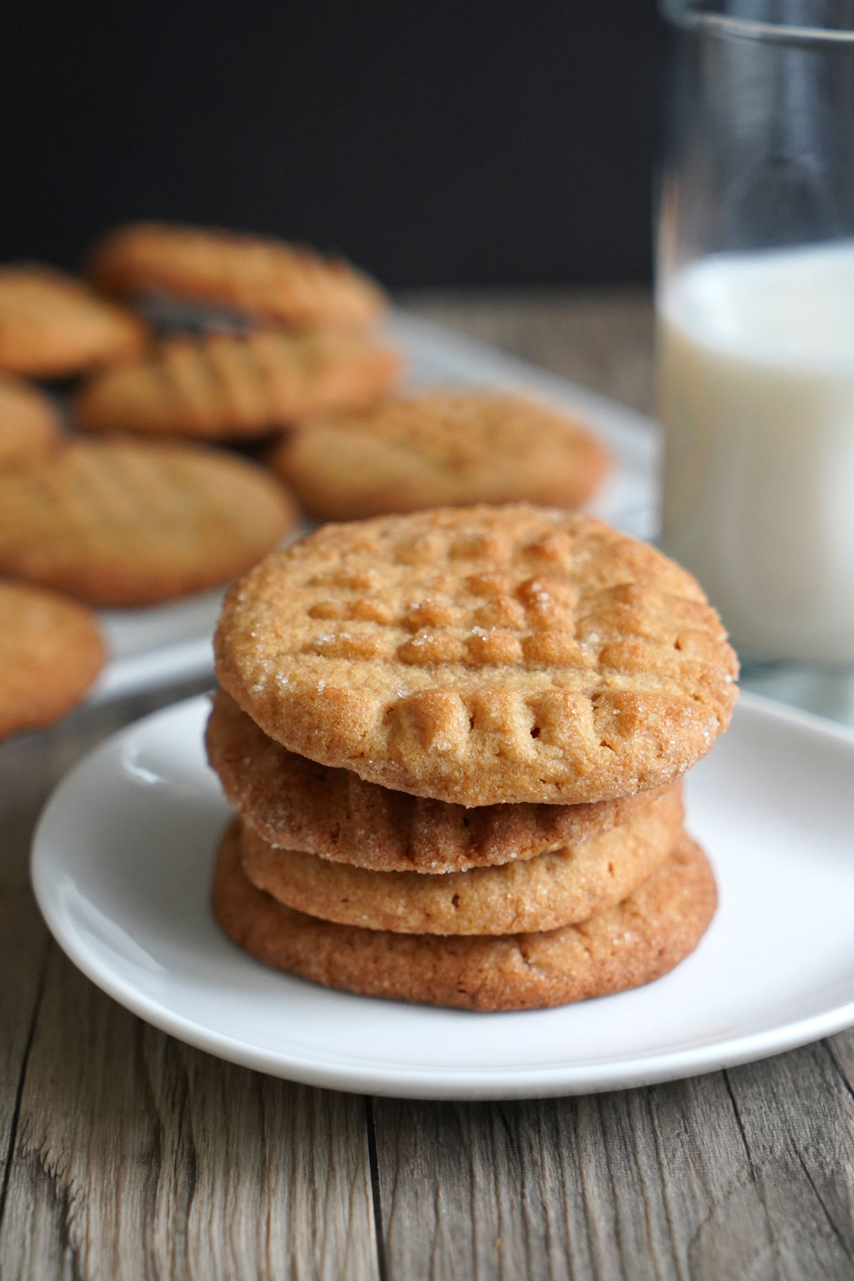 Stacked peanut butter cookies in awhite plate with milk in background.