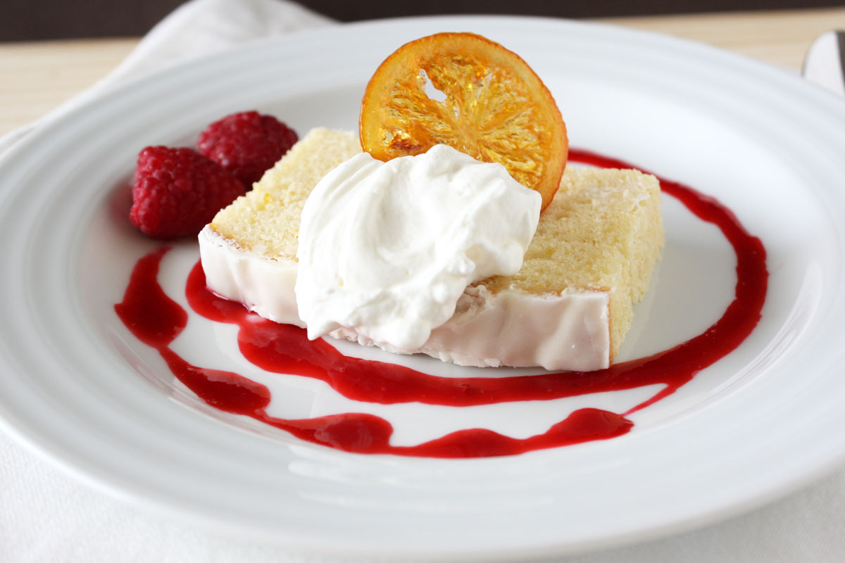 Slice of pound cake in a white plate surrounded by raspberry sauce and topped with whipped cream and candied lemon.