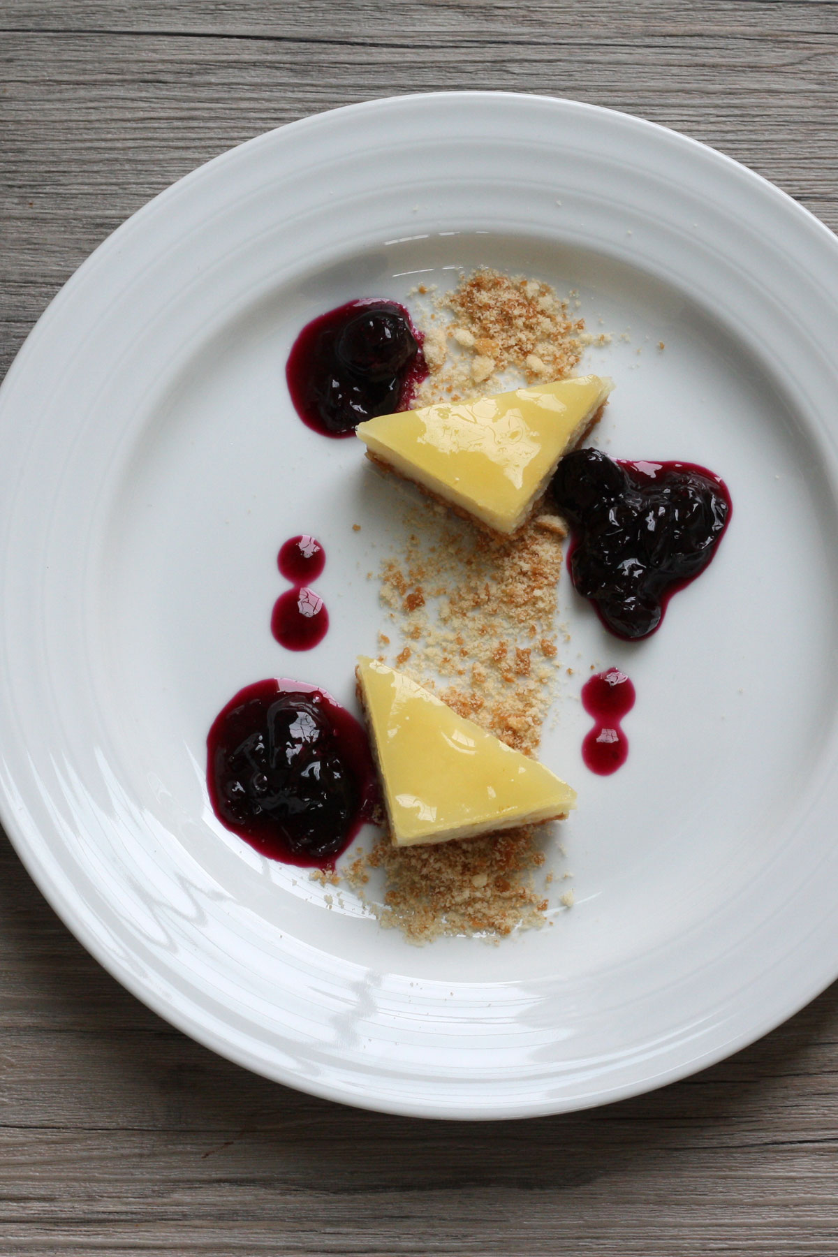 Meyer lemon cheesecake bar cut into two triangles and plated with blueberry compote and crushed graham cracker. 
