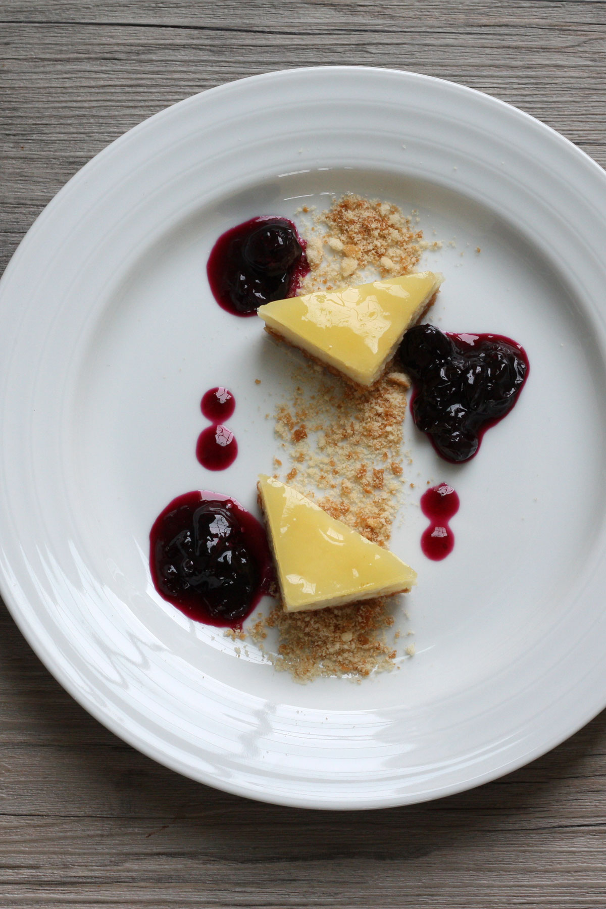 Meyer lemon cheesecake bar cut into two triangles and plated with blueberry compote and crushed graham cracker.