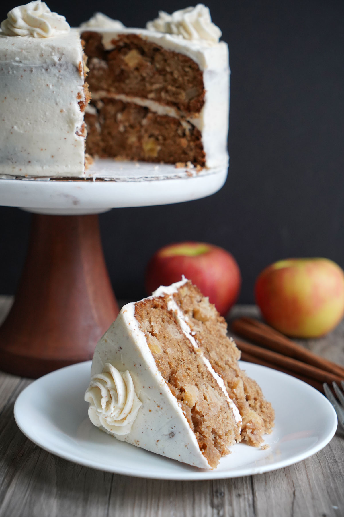 Slice of apple spice cake on a white plate with cake on cake stand in background.