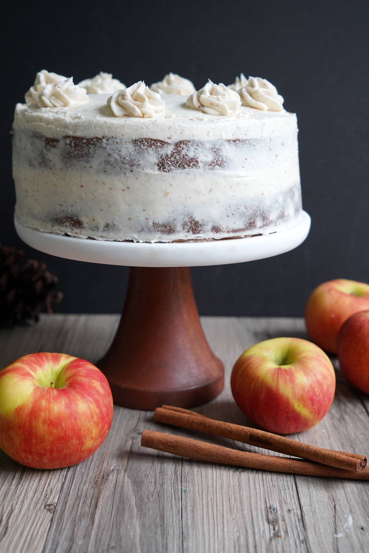 Frosted apple spice cake on a cake stand with apples and cinnamon surrounding the stand.