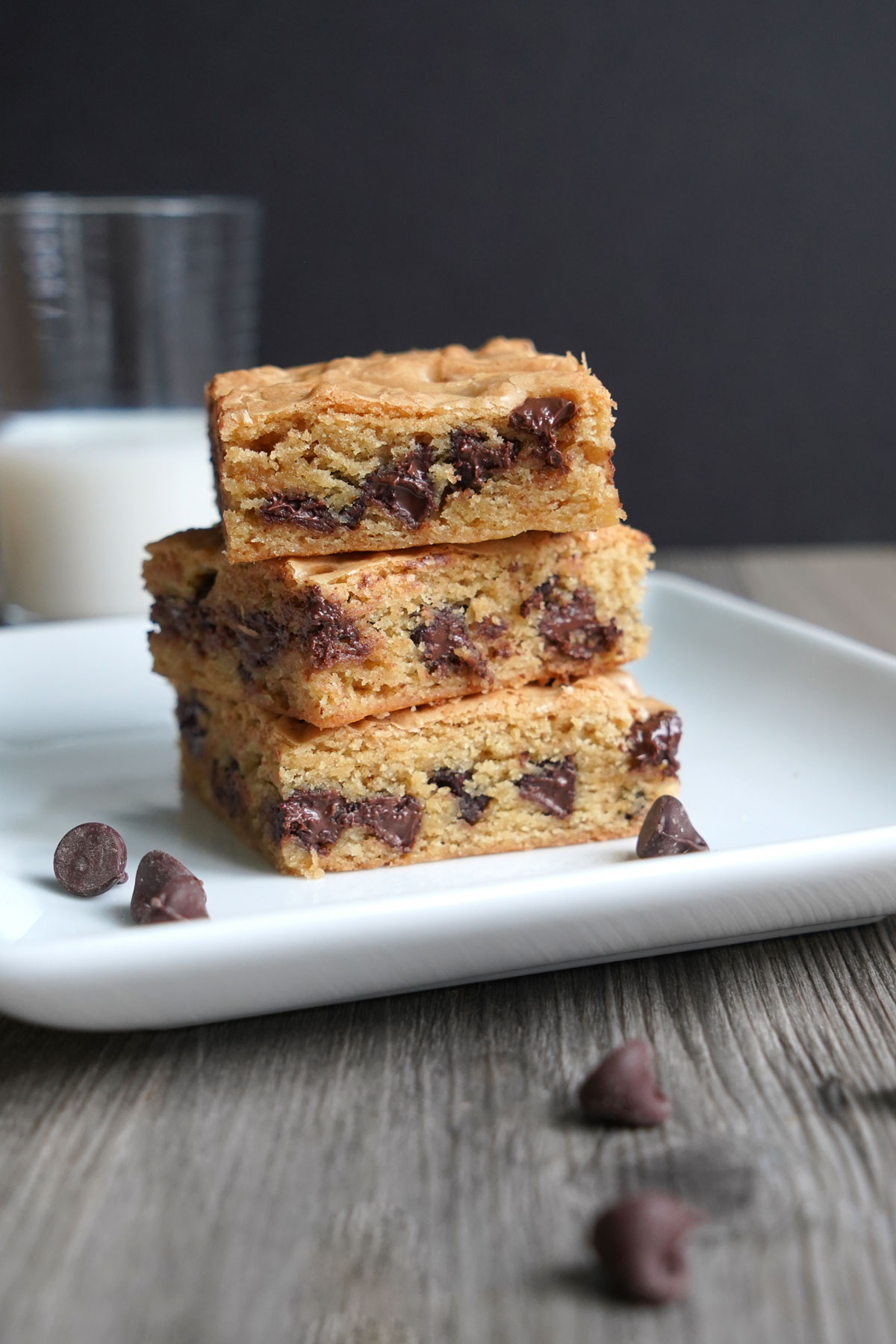 Three chocolate chip cookie bars stacked on a plate with milk glass in background.
