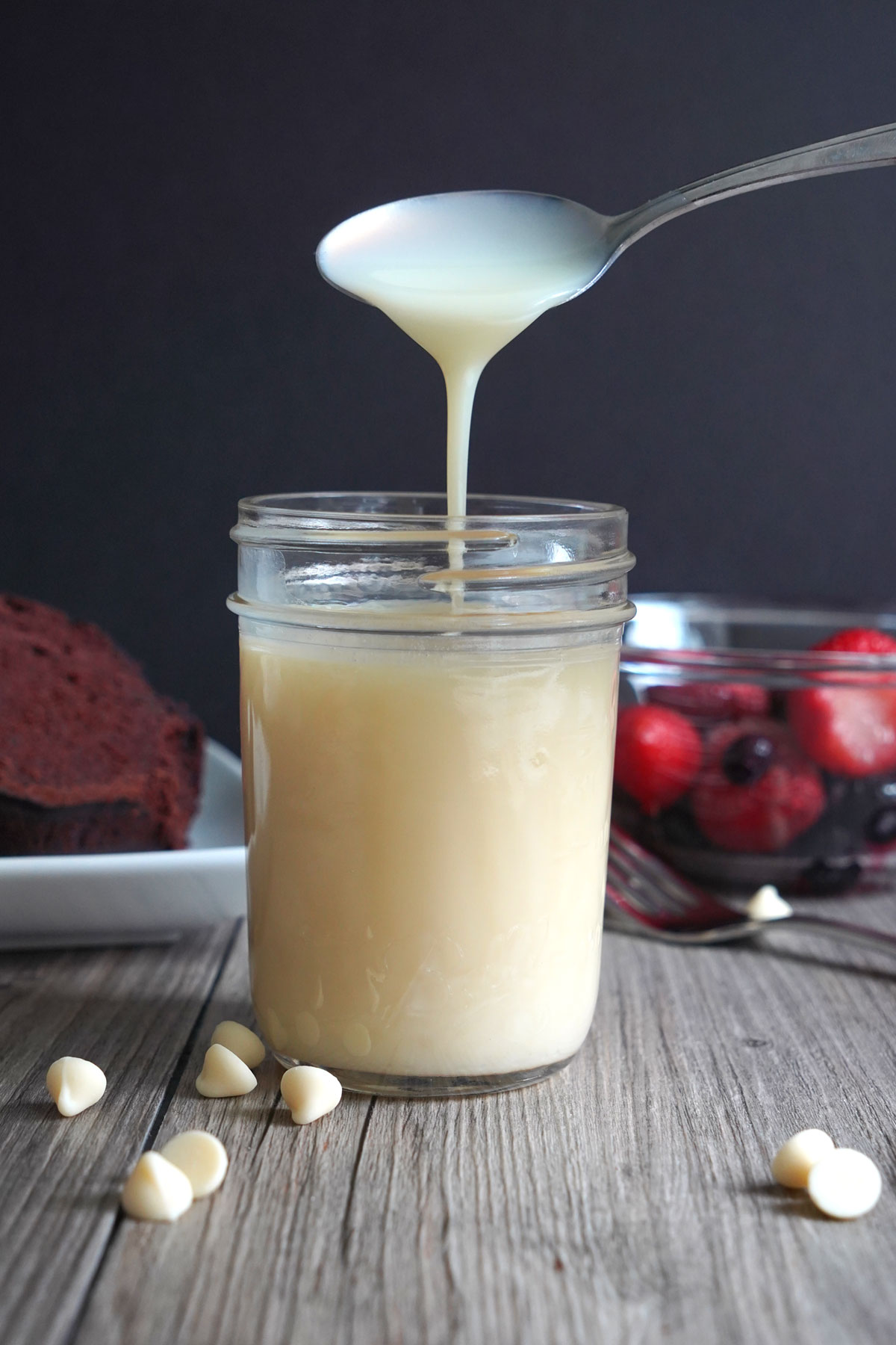 White chocolate sauce in a glass jar with spoon dripping sauce back into jar to show consistency.