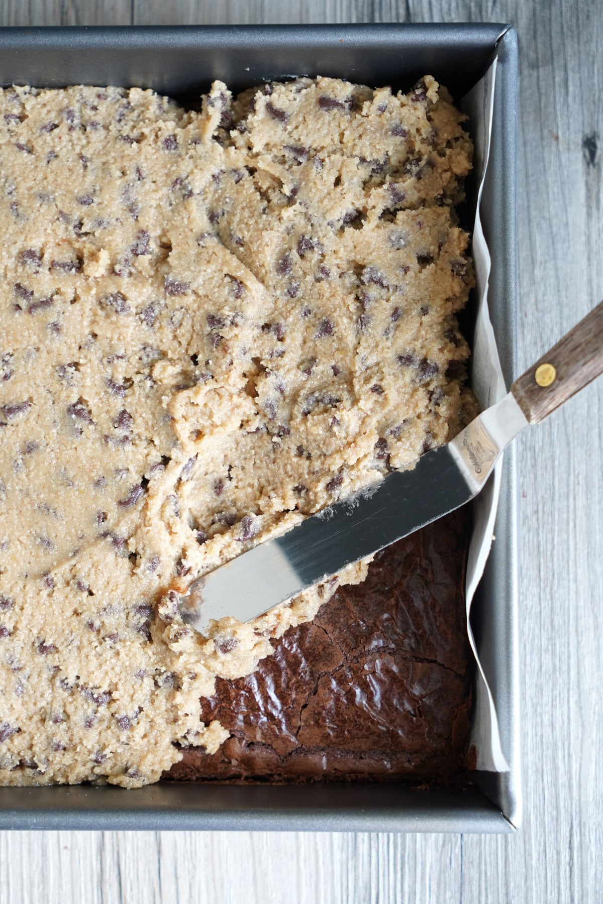 Offset spatula spreading a thick layer of edible cookie dough frosting over cooled brownies in a pan