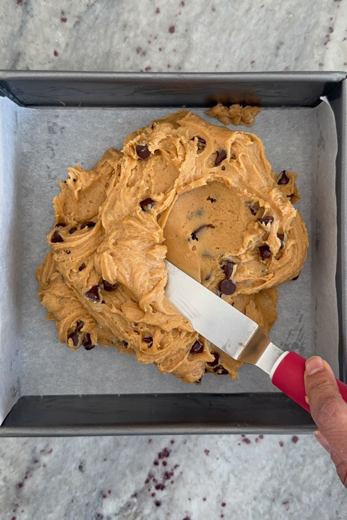 Peanut butter bar batter being spread into 8x8 inch pan with offset spatula.