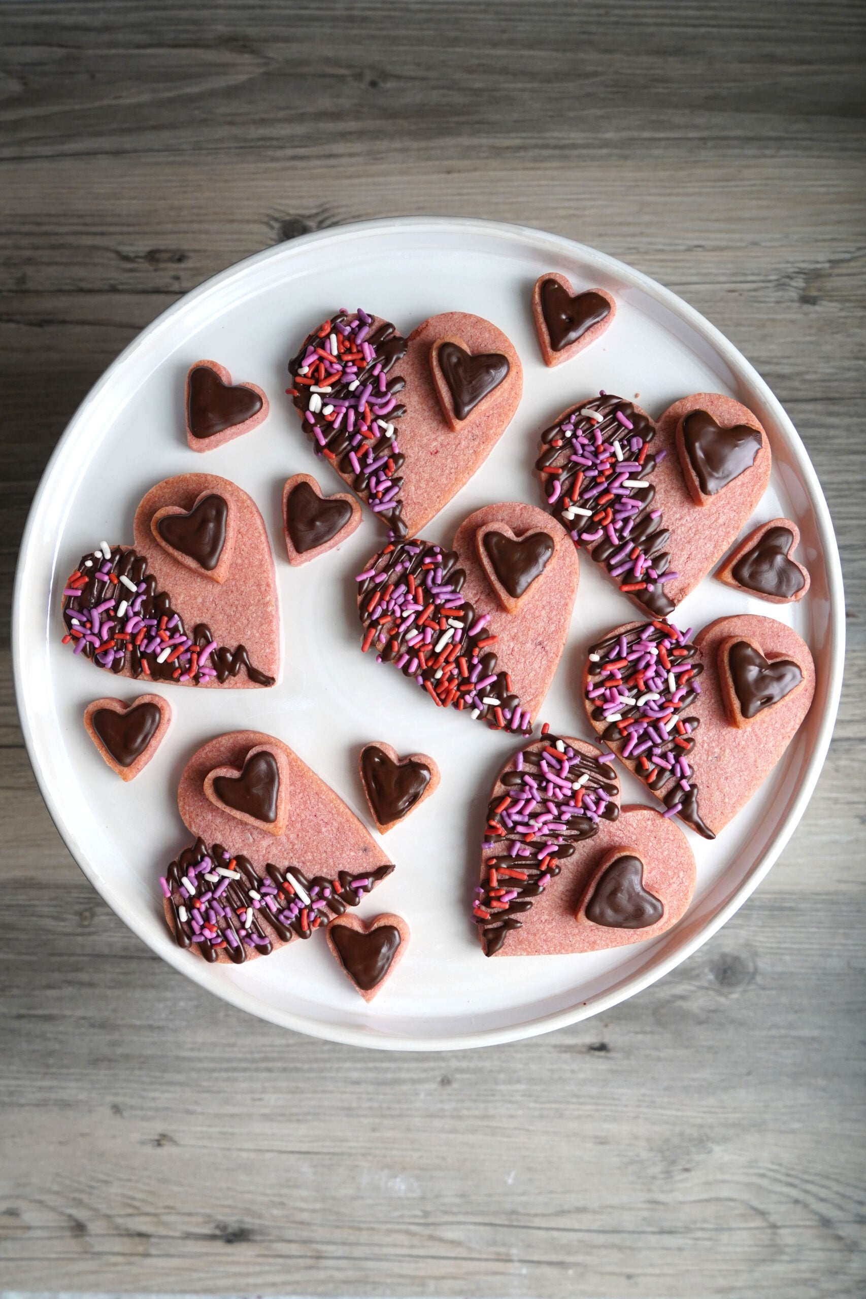 Heart shaped cookies in two sizes made with pink dough and decorated with chocolate and sprinkles on a cake stand.