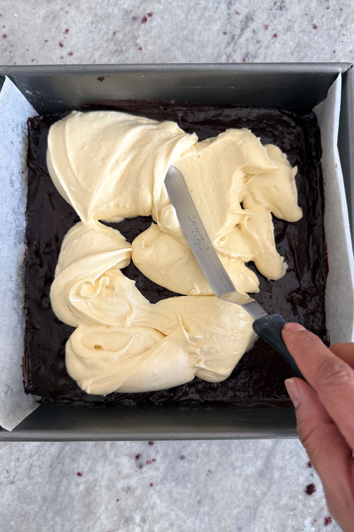 Cheesecake batter being spread over brownie batter.