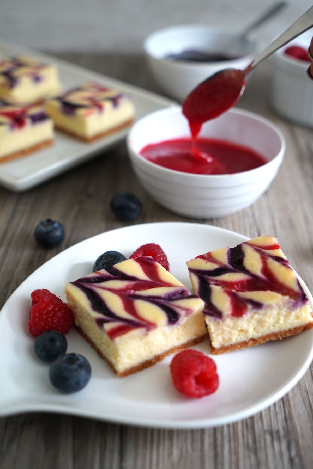 Red white blue cheesecake bars on a plate. Raspberry sauce in background being spooned out to drizzle over cheesecake.