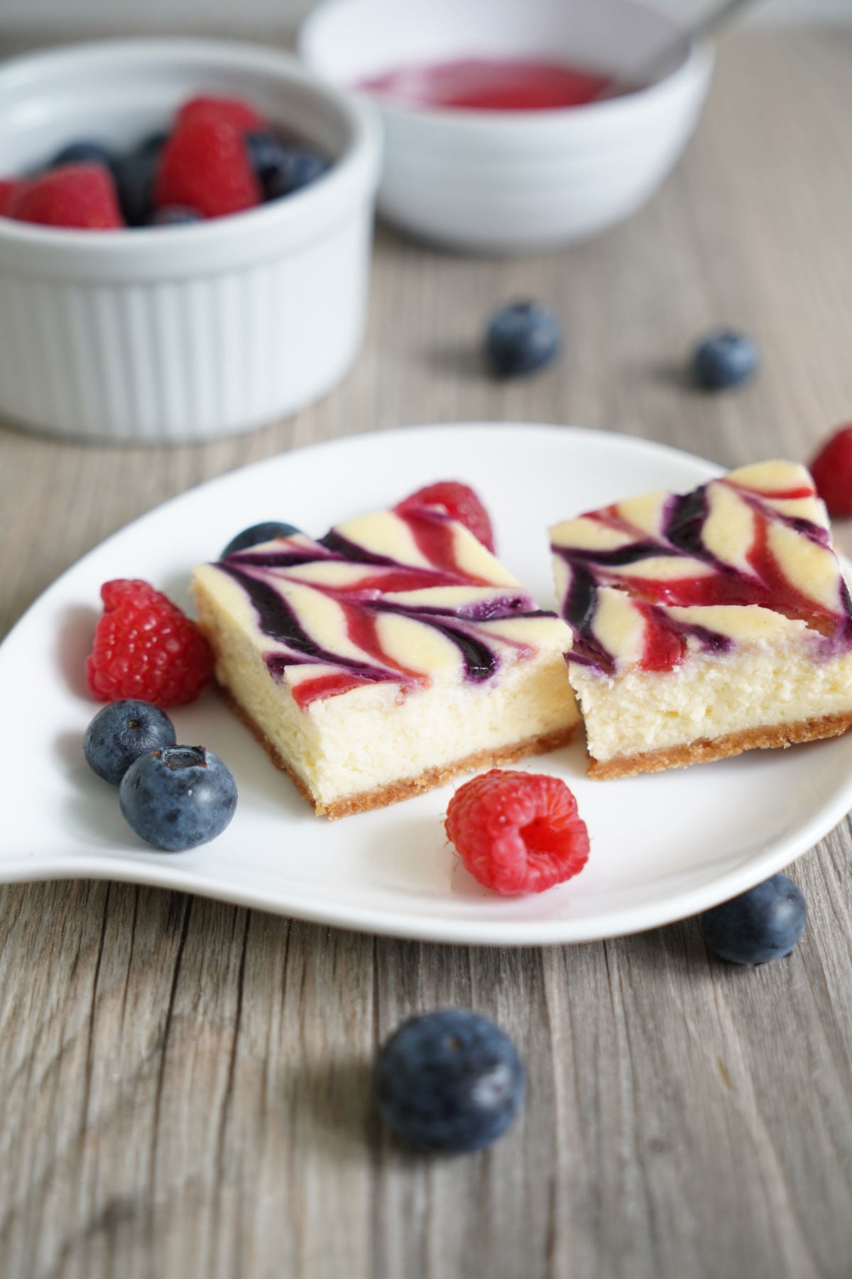 Two cheesecake bars in white plate with berries scattered around.