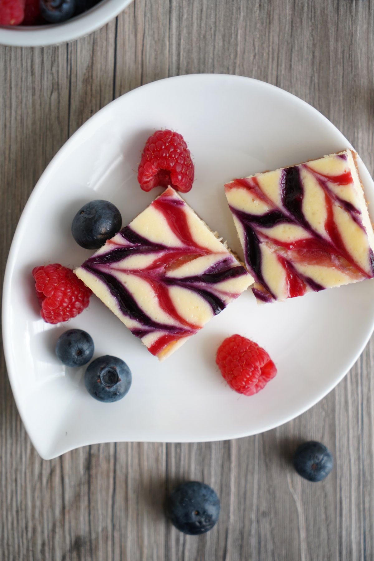 Top view: Two cheesecake bars in white plate with berries scattered around.