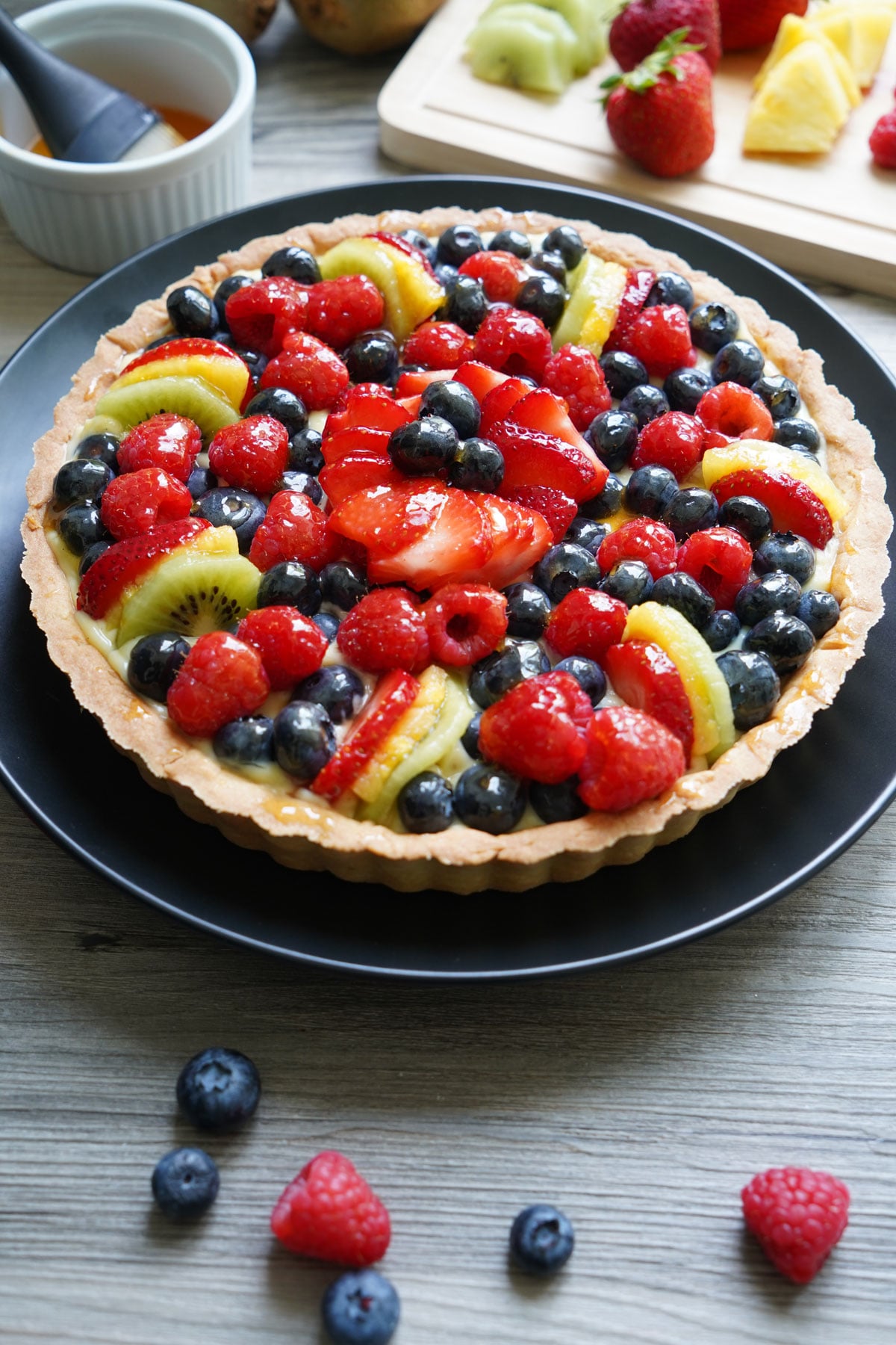 Fruit tart on a black plate with cut fruit in the background.