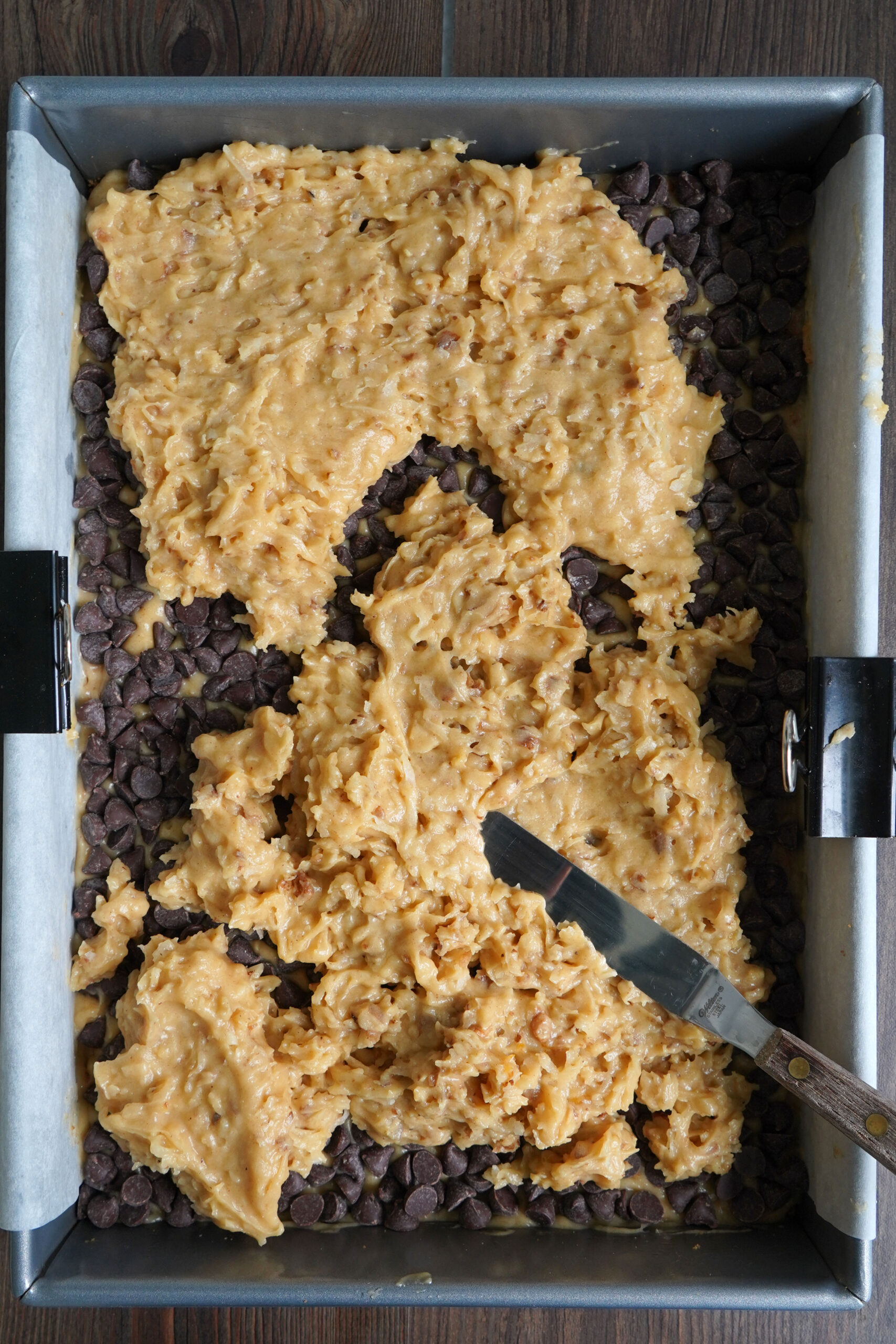Paradise pie's coconut and walnut layer being spread over chocolate chip layer in a 9x13 pan.