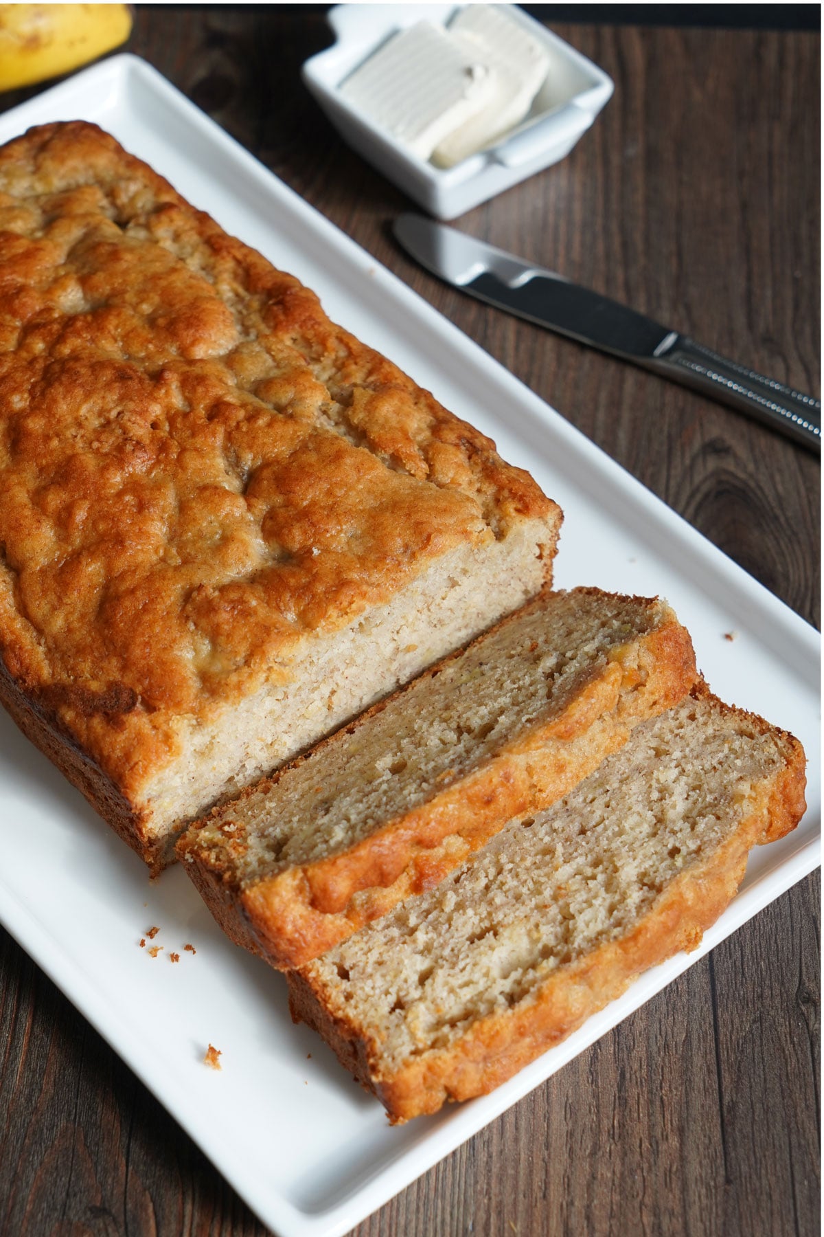 Overhead view of pancake mix banana bread loaf showing the top color and crumb on slices. Pats of butter for serving on the side.