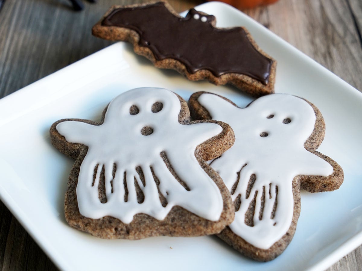 Ghost sugar cookies flooded with white icing, with a black icing bat behind them.