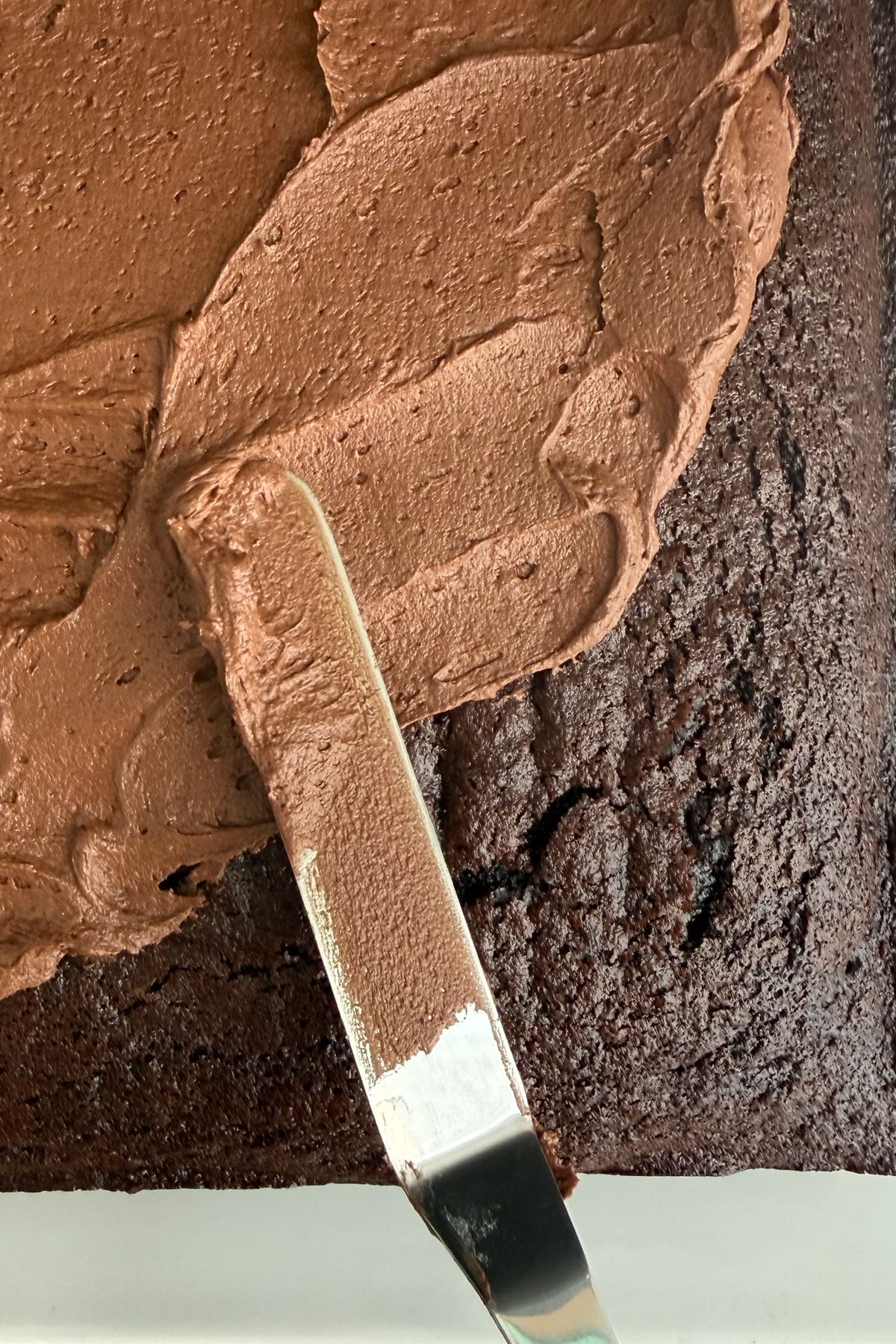 Fudge frosting being spread over the top of a cooled chocolate cake.